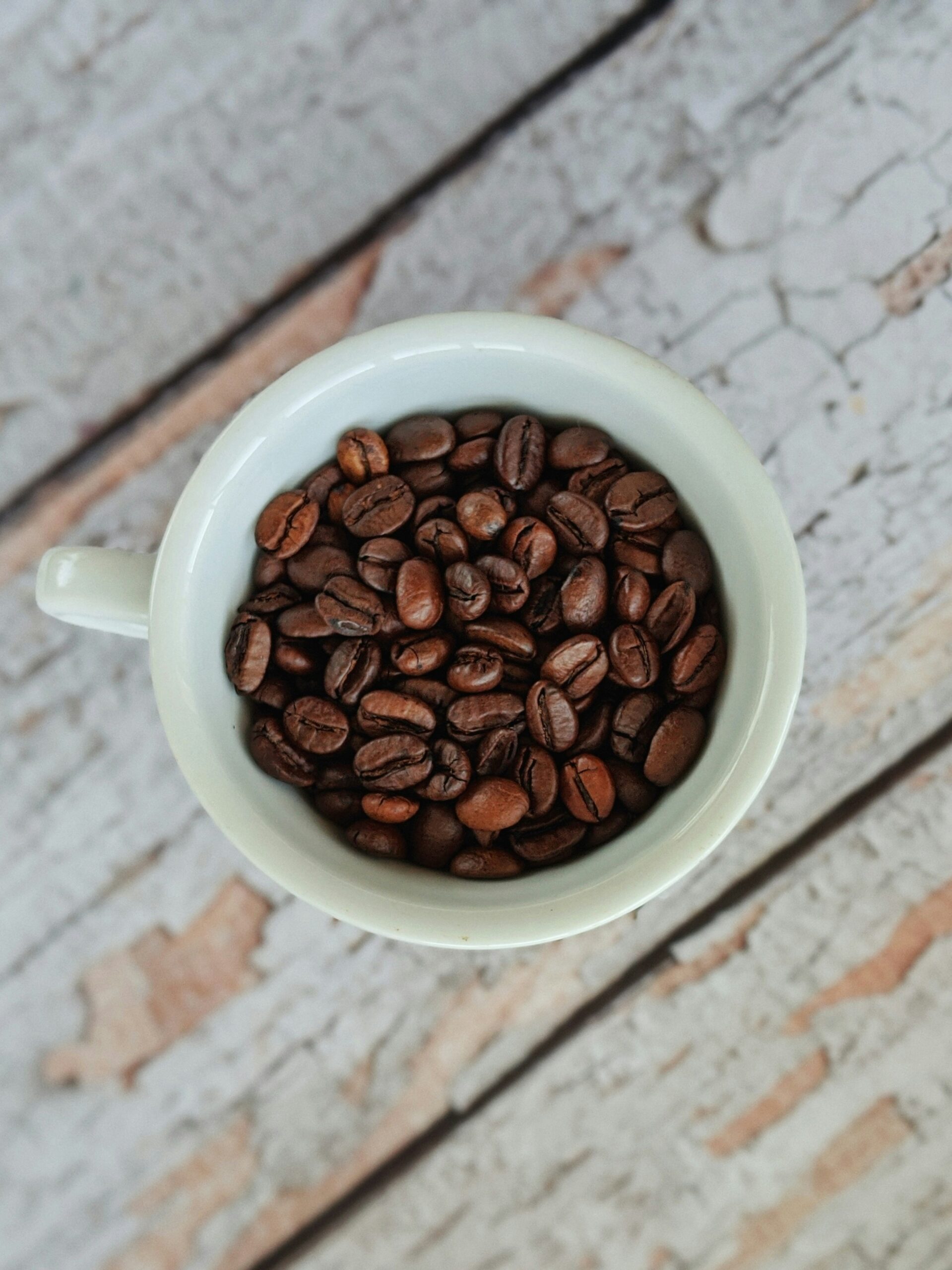 Overhead view of dark roast coffee beans filling a white ceramic cup with handle, positioned on a whitewashed wooden surface