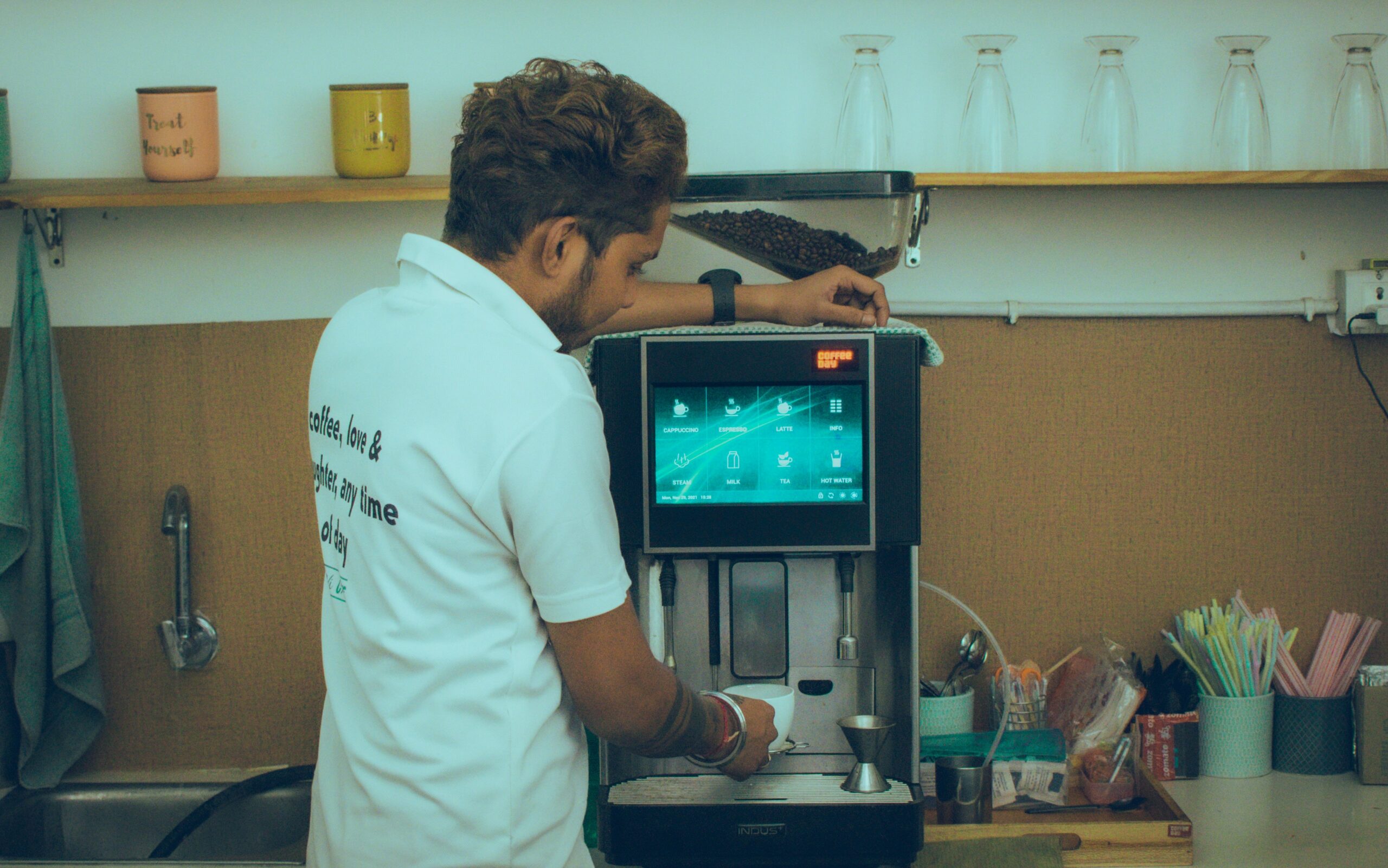 A staff member in a white t-shirt topping up the bean hopper on a commercial bean-to-cup machine with a touchscreen display in a café setting.