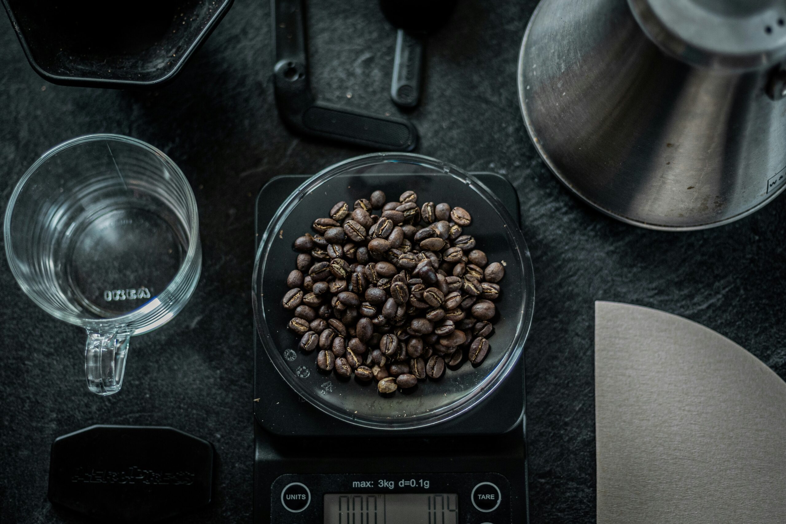 Overhead view of coffee beans in a glass bowl on a digital scale showing, surrounded by a glass of water, metal measuring cups, and metal brewing equipment on a dark surface