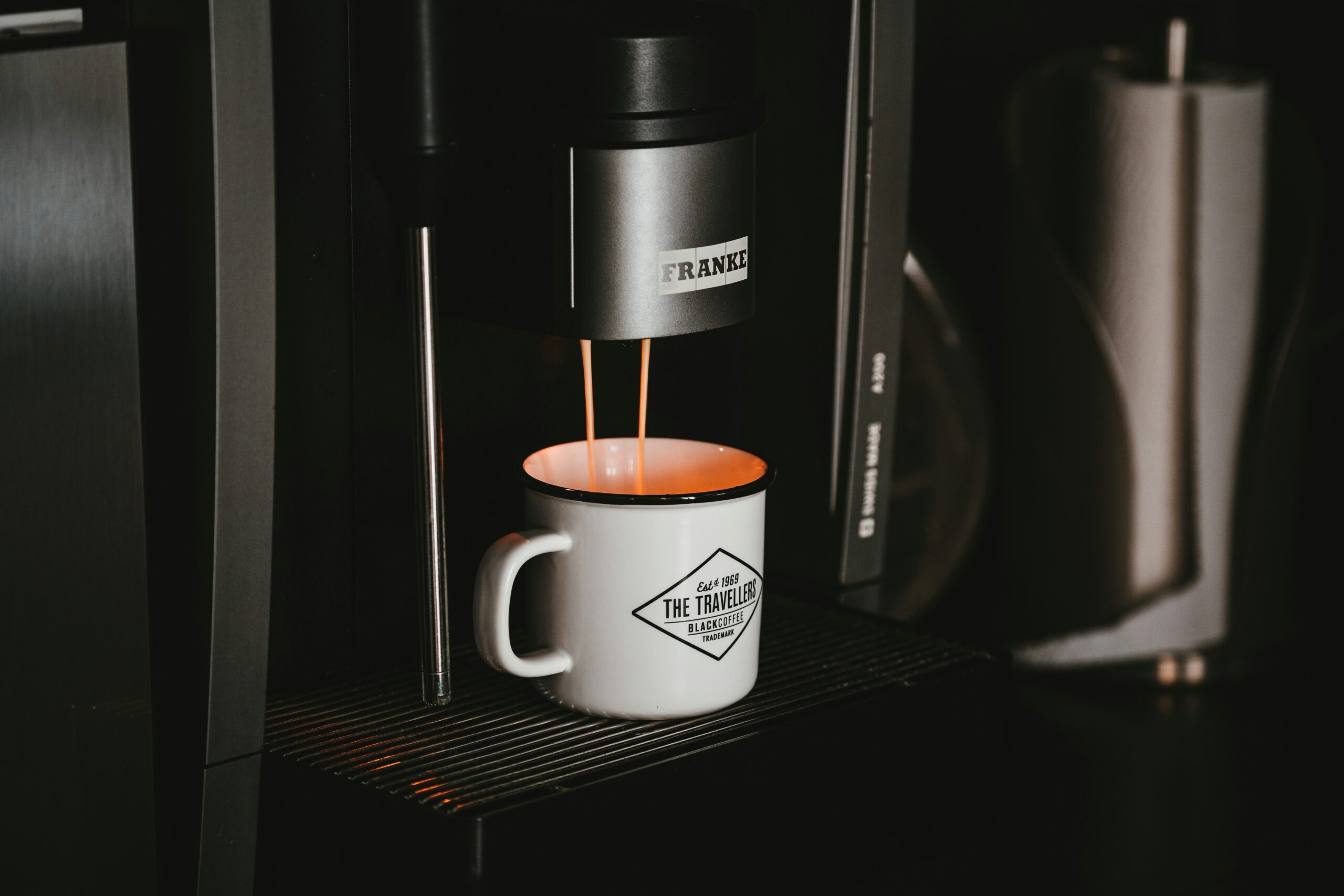 A Franke bean-to-cup coffee machine dispensing fresh coffee into a branded enamel mug, against a dark background.