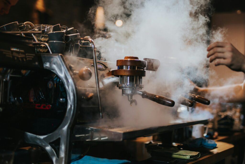 Close-up of an espresso machine steaming, with a portafilter attached and a barista nearby.