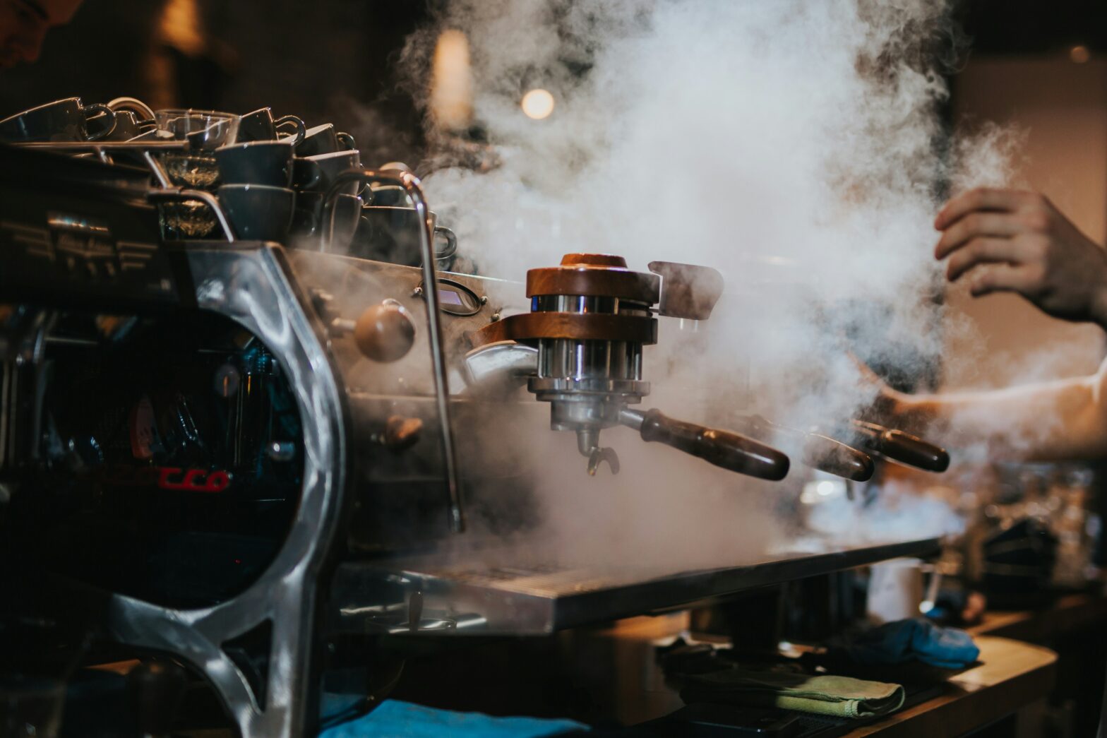 espresso machine steaming blog hero - Indigo Valley Close-up of an espresso machine steaming, with a portafilter attached and a barista nearby.