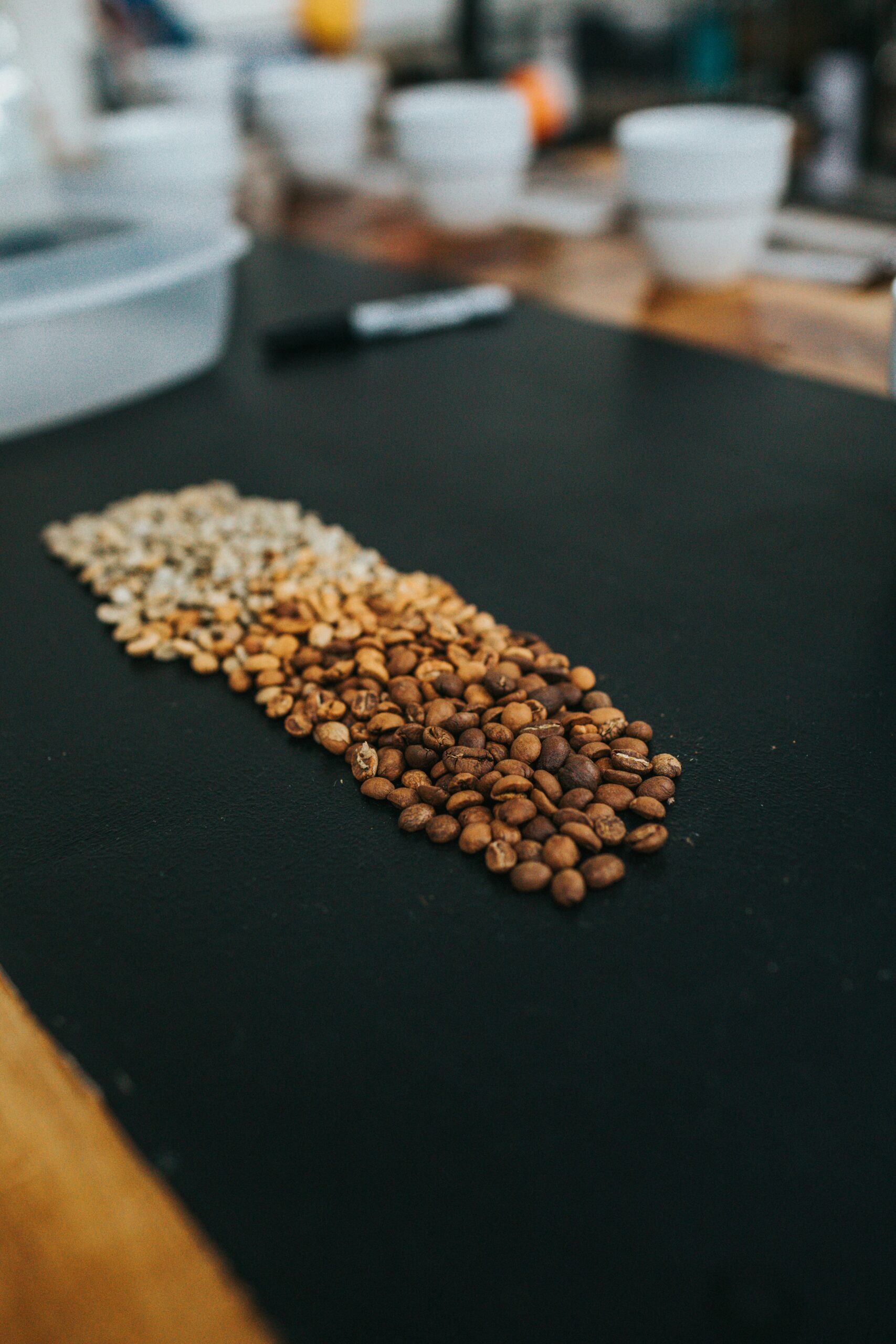 Coffee bean roast progression displayed on a dark surface, showing gradual color change from light green-tan beans on the left through medium brown to dark brown beans on the right, with white cups and tasting supplies blurred in the background