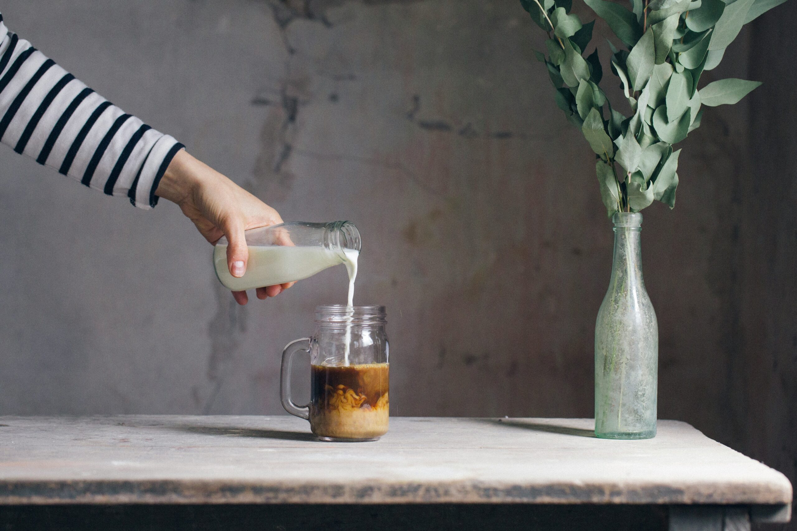 A person in a striped top pouring milk from a glass bottle into a mason jar of coffee on a rustic table, beside a clear bottle of eucalyptus stems.