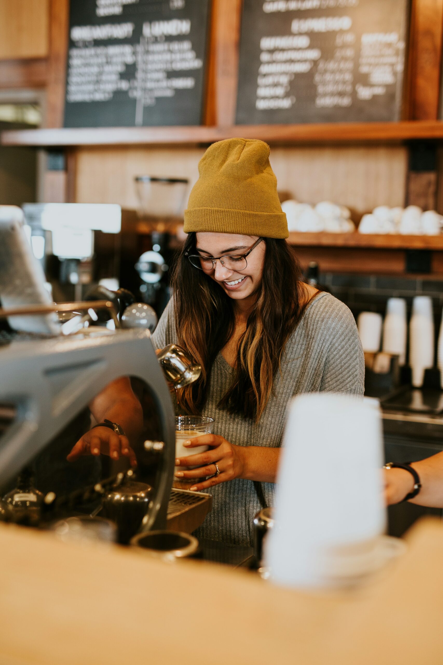 brooke-cagle-9fHMo1-5Io8-unsplash - Indigo Valley A smiling barista in a mustard beanie and glasses pouring steamed milk into a cup at an espresso machine, with menu boards behind.