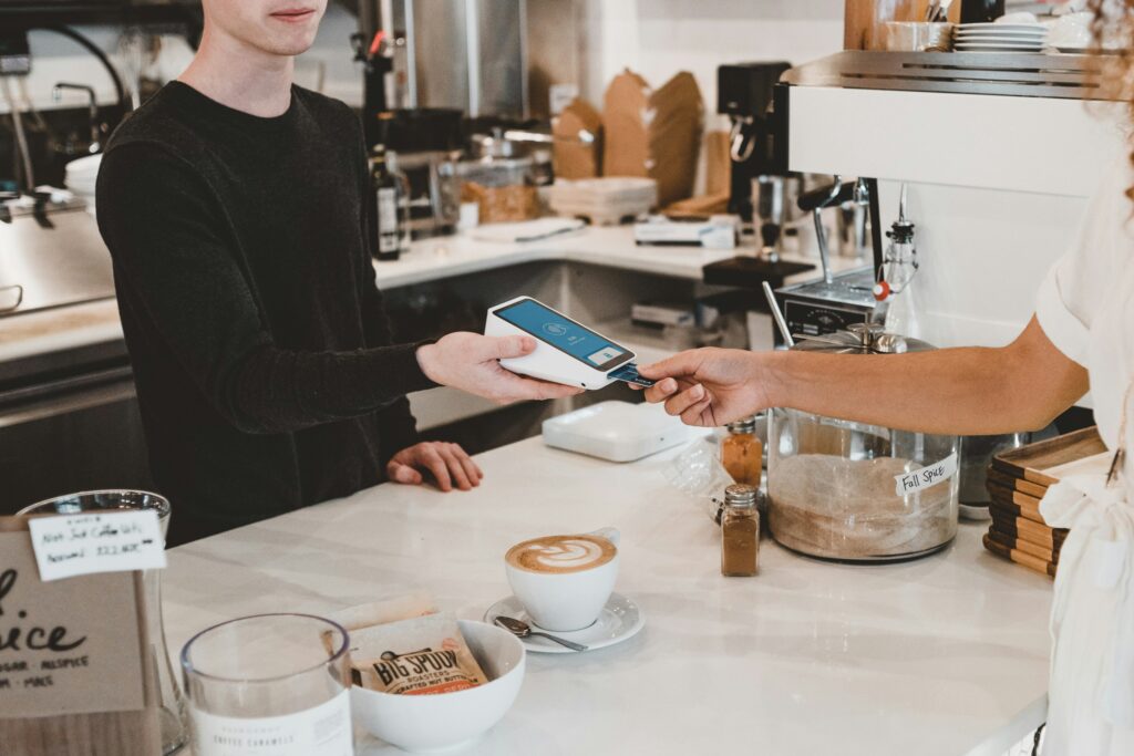 clay-banks-oNm9NkTFLfA-unsplash - Indigo Valley A customer paying by card at a cafe counter as the barista holds out a card reader, with a latte and pastries on the white countertop.