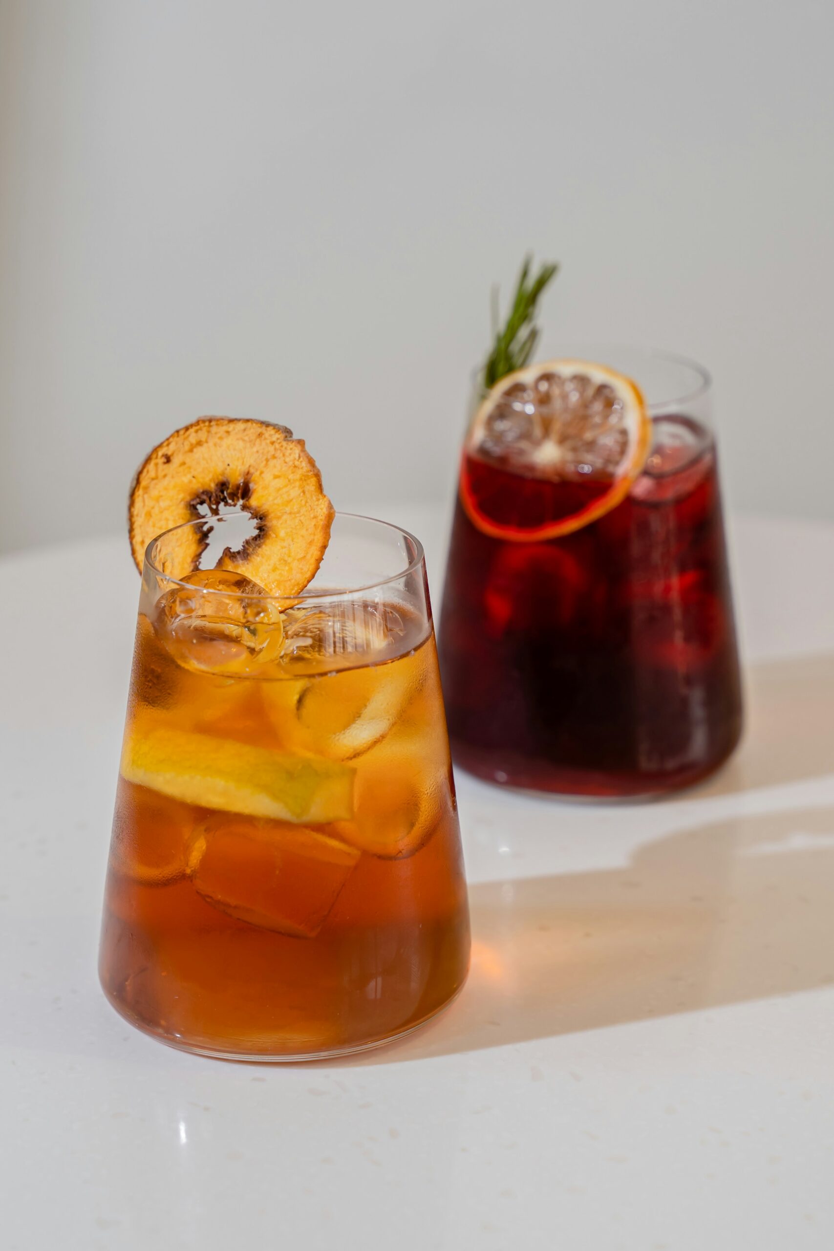 Two glasses of iced fruit tea on a white surface, garnished with dried orange slices, lemon and a sprig of rosemary - one amber, one deep red.