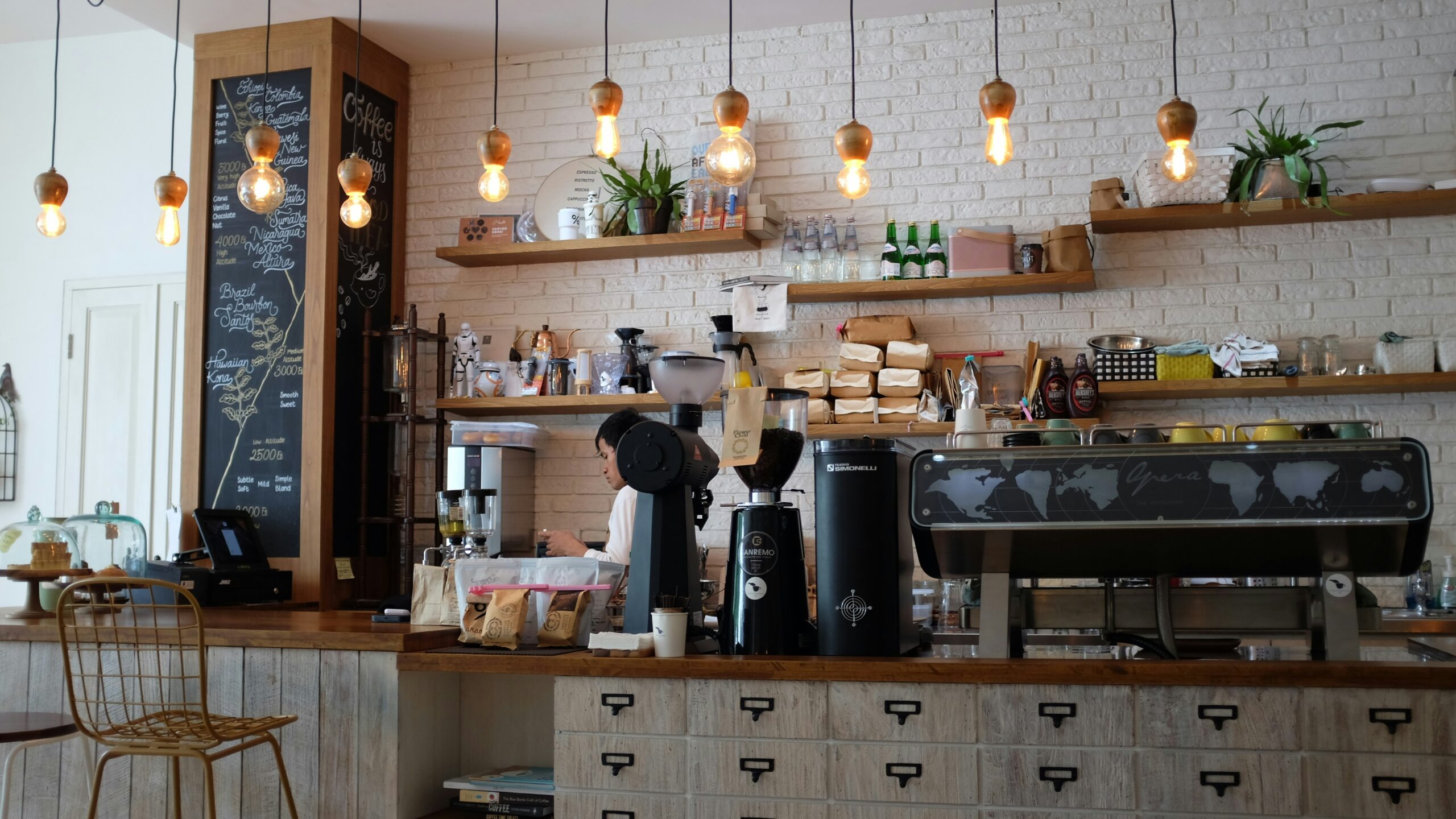 nafinia-putra-Kwdp-0pok-I-unsplash - Indigo Valley Interior of a cafe with a barista behind a wooden counter, a commercial espresso machine, bags of coffee beans and a chalkboard listing coffee origins.