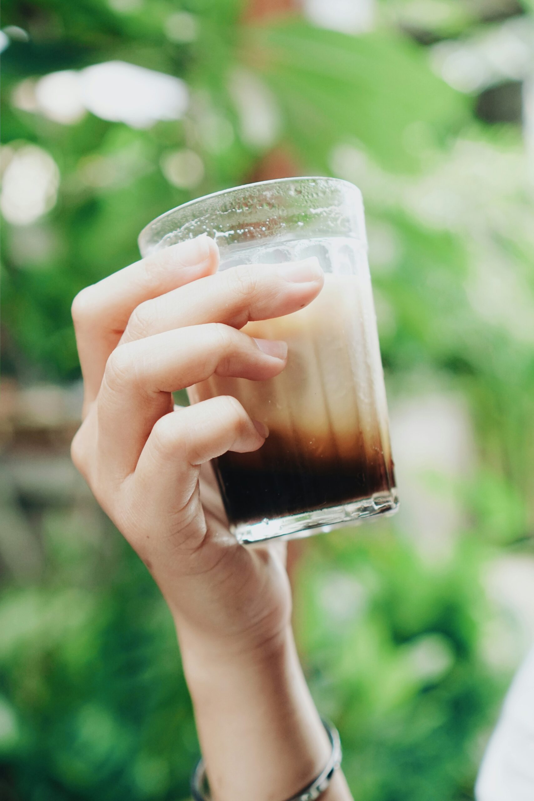 A hand raising a tall glass of layered iced coffee outdoors, surrounded by green tropical foliage.
