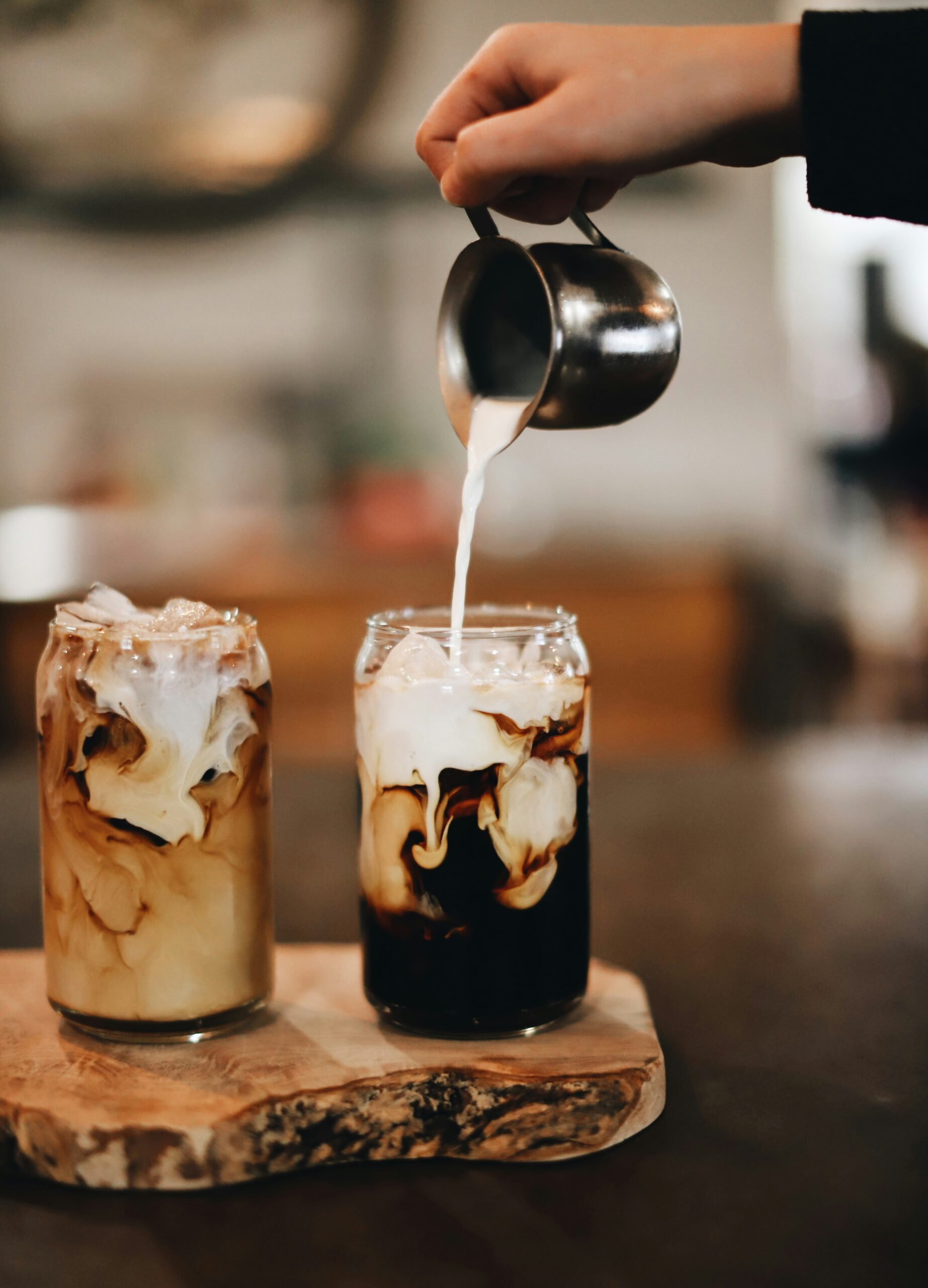 Two glasses of iced coffee on a rustic wooden board, with milk being poured from a small metal jug and swirling through the dark coffee.