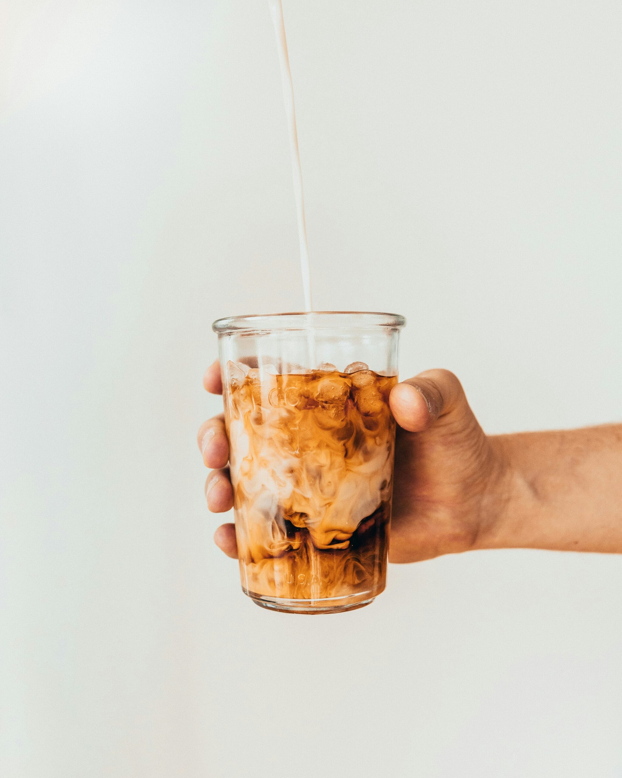 A hand holding a tall glass of iced latte against a plain pale background as milk is poured in, marbling through the ice.