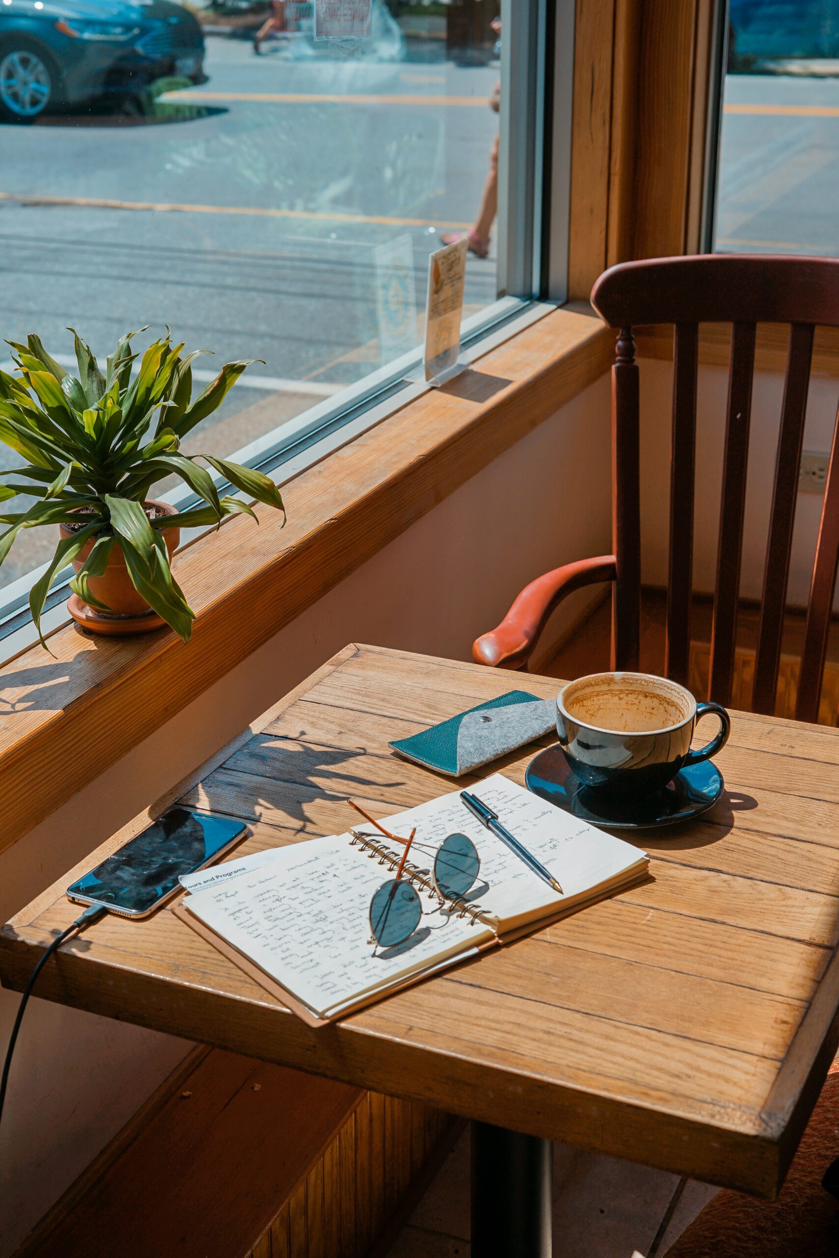 toa-heftiba-QnUywvDdI1o-unsplash - Indigo Valley A cup of coffee on a sunlit cafe window table beside an open notebook, pen and sunglasses, with a phone charging and a potted plant on the windowsill.