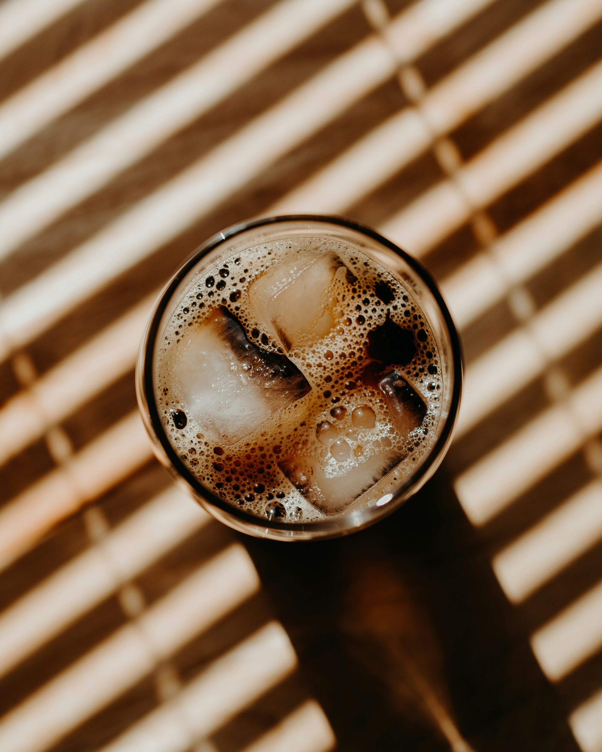 Overhead view of a glass of iced cold brew coffee topped with ice cubes and fine foam, on a wooden surface striped with sunlight through blinds.