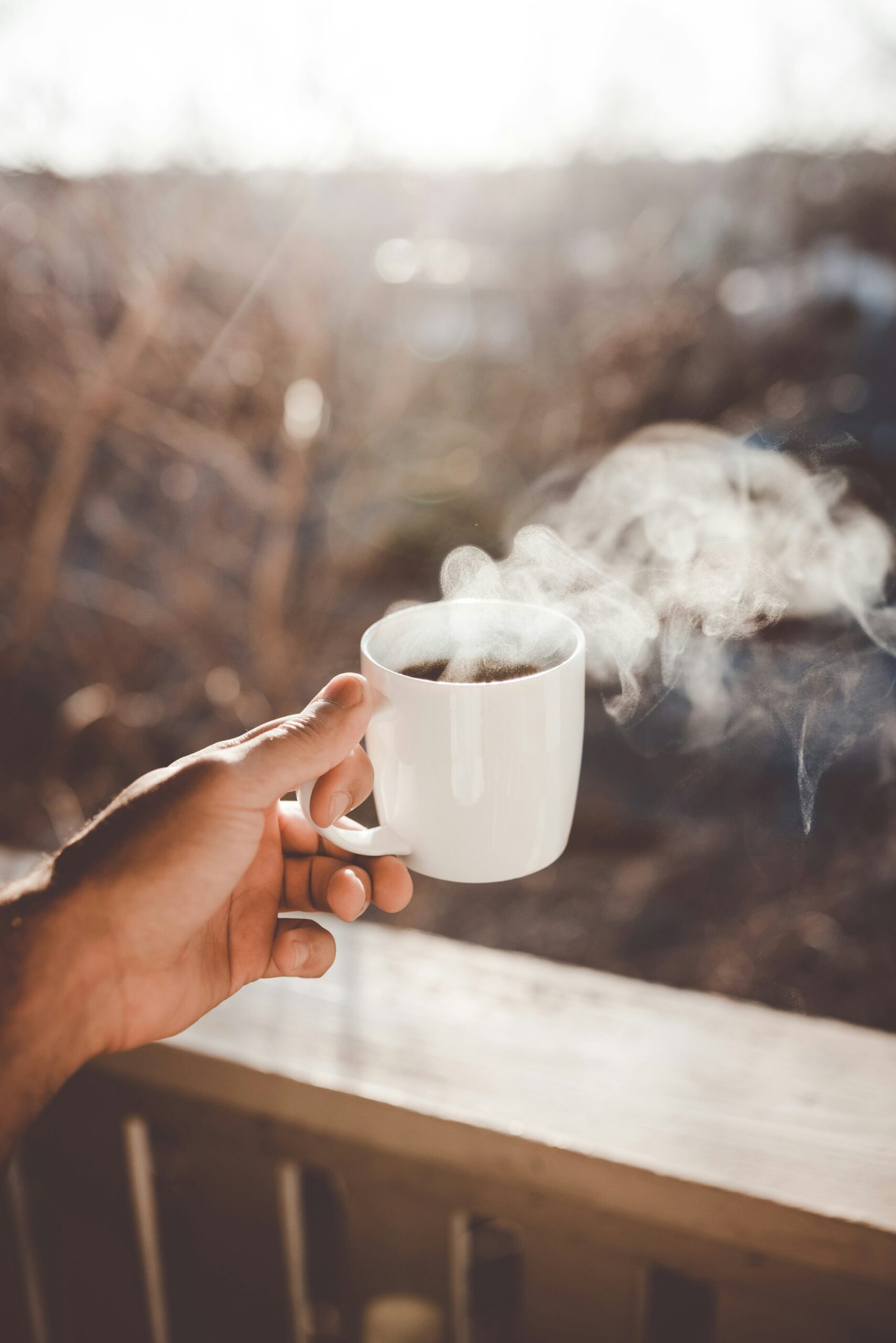 clay-banks-_wkd7XBRfU4-unsplash - Indigo Valley A hand holding a white mug of steaming black coffee outdoors, backlit by morning sunlight over a wooden railing.
