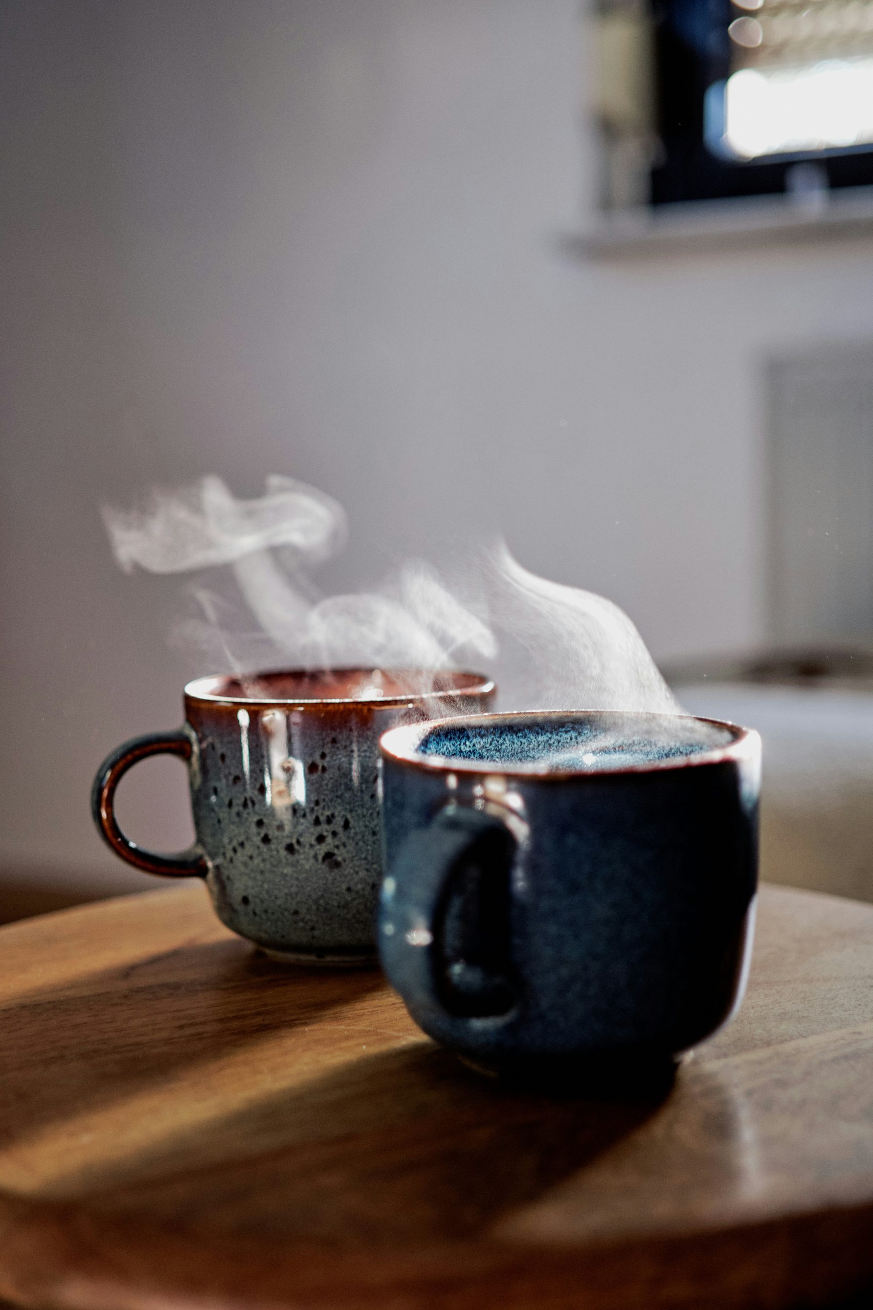 florian-siedl-nfr8Miay5zw-unsplash - Indigo Valley Two rustic speckled ceramic mugs of steaming coffee side by side on a wooden tabletop in soft natural light.