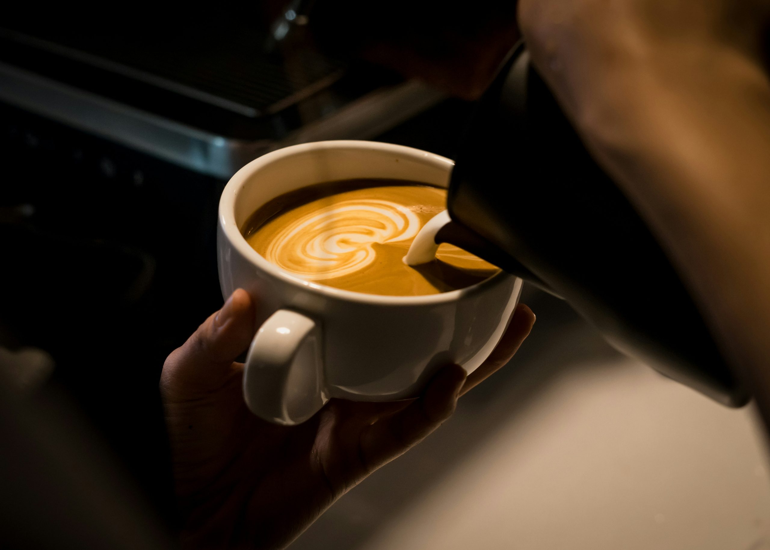 jayden-sim-pQmlo0juG50-unsplash - Indigo Valley A hand holding a cup of coffee topped with delicate leaf latte art, in warm, moody lighting.