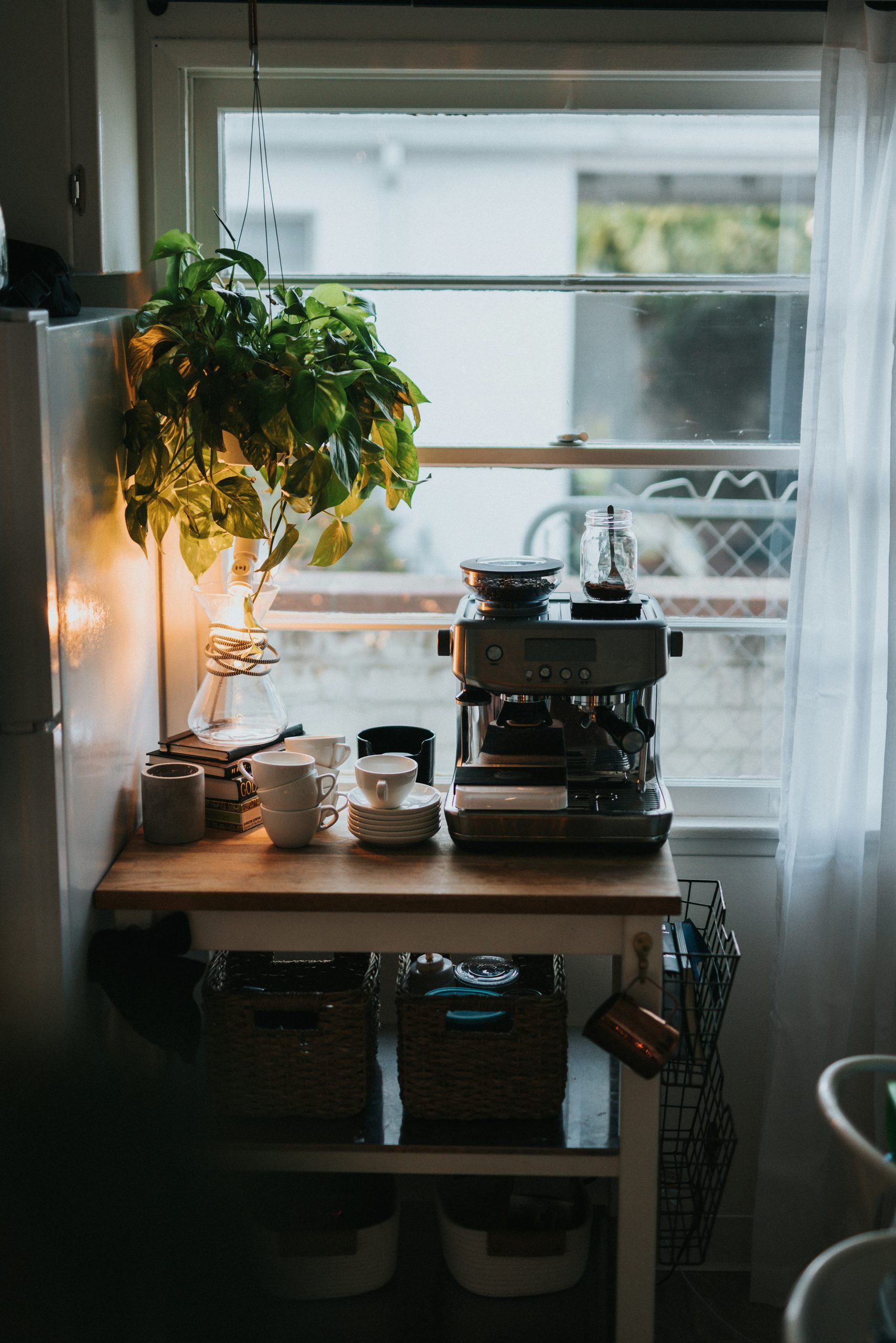 nathan-dumlao-ioVRpz0gX5w-unsplash - Indigo Valley A cosy home coffee station by a window, with an espresso machine on a wooden shelf, stacked mugs and a trailing potted plant lit by warm daylight.