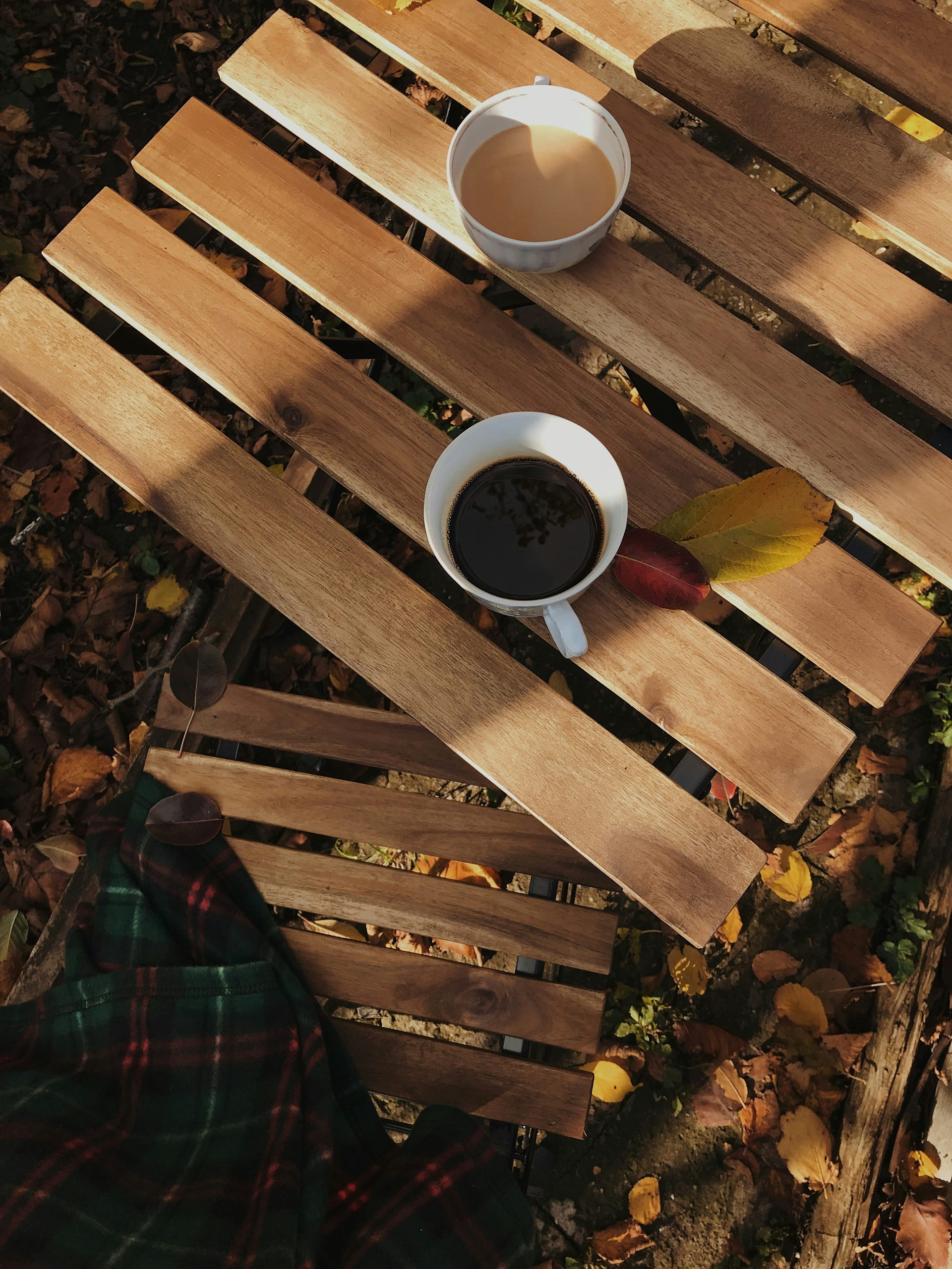 olly-hoodyakova-8VSmseBs-o0-unsplash - Indigo Valley An overhead view of two cups of black coffee on weathered wooden planks, surrounded by fallen autumn leaves.