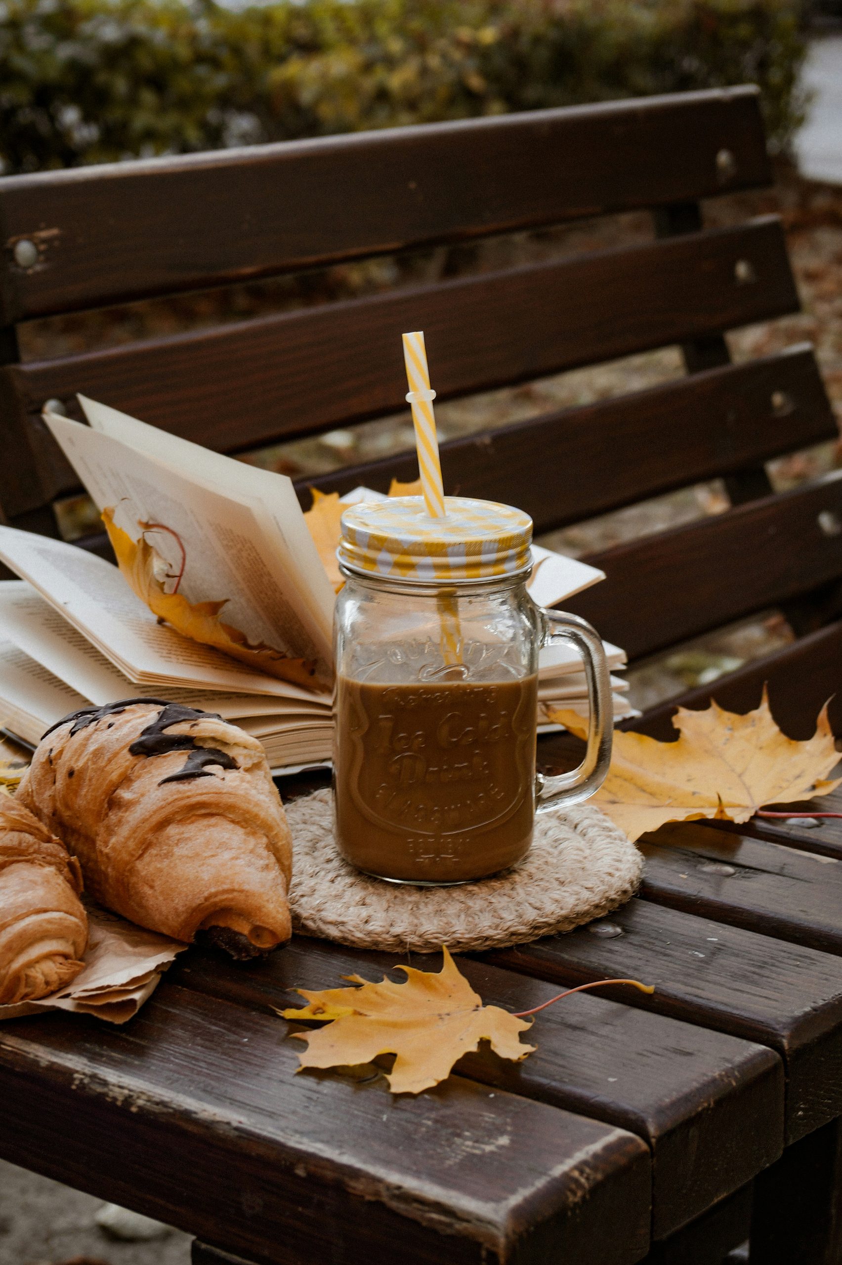 valentina-ivanova-pgEvcMI7M_g-unsplash - Indigo Valley A glass mason jar mug of coffee with a paper straw on a round coaster, with a croissant and scattered autumn leaves on a dark wooden bench.